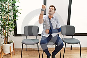 Handsome young man sitting at doctor waiting room with arm injury angry and mad raising fist frustrated and furious while shouting