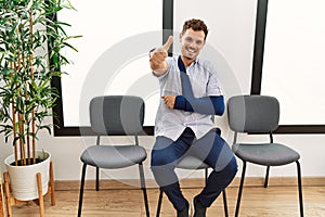 Handsome young man sitting at doctor waiting room with arm injury approving doing positive gesture with hand, thumbs up smiling
