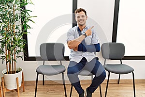 Handsome young man sitting at doctor waiting room with arm injury with a big smile on face, pointing with hand finger to the side