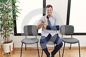 Handsome young man sitting at doctor waiting room with arm injury doing happy thumbs up gesture with hand