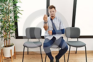 Handsome young man sitting at doctor waiting room with arm injury doing italian gesture with hand and fingers confident expression