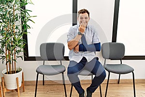 Handsome young man sitting at doctor waiting room with arm injury laughing and embarrassed giggle covering mouth with hands,