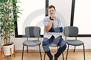 Handsome young man sitting at doctor waiting room with arm injury looking confident at the camera smiling with crossed arms and