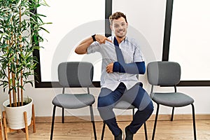 Handsome young man sitting at doctor waiting room with arm injury looking confident with smile on face, pointing oneself with