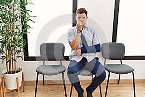 Handsome young man sitting at doctor waiting room with arm injury looking stressed and nervous with hands on mouth biting nails