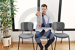 Handsome young man sitting at doctor waiting room with arm injury looking unhappy and angry showing rejection and negative with