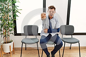 Handsome young man sitting at doctor waiting room with arm injury pointing displeased and frustrated to the camera, angry and