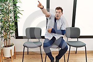 Handsome young man sitting at doctor waiting room with arm injury pointing with finger surprised ahead, open mouth amazed