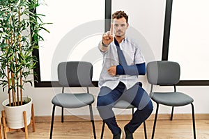 Handsome young man sitting at doctor waiting room with arm injury pointing with finger up and angry expression, showing no gesture