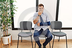 Handsome young man sitting at doctor waiting room with arm injury shocked covering mouth with hands for mistake