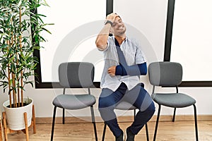 Handsome young man sitting at doctor waiting room with arm injury smiling confident touching hair with hand up gesture, posing