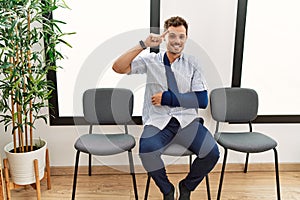 Handsome young man sitting at doctor waiting room with arm injury smiling pointing to head with one finger, great idea or thought,