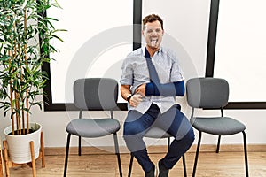 Handsome young man sitting at doctor waiting room with arm injury sticking tongue out happy with funny expression