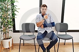 Handsome young man sitting at doctor waiting room with arm injury surprised pointing with finger to the side, open mouth amazed