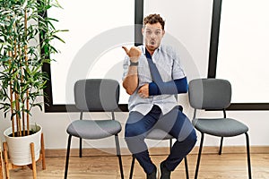 Handsome young man sitting at doctor waiting room with arm injury surprised pointing with hand finger to the side, open mouth