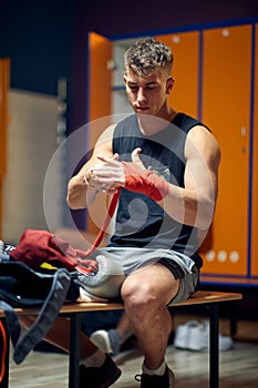 Handsome young man sitting in dressing room, focusing while tying tape around his hand for protection