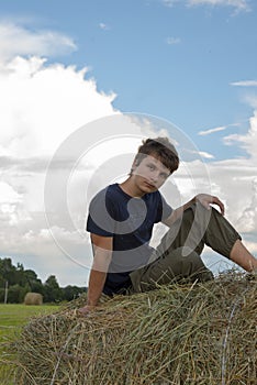 A handsome young man is sitting on a haystack against a blue cloudy sky