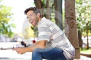 Handsome young man sitting outdoors with mobile phone and laughing