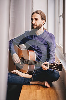 Handsome young man sitting on the windowsill and playing acoustic guitar