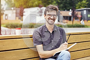 Handsome young man smiling face writing in a note book sitting outside. Guy wearing glasses alone working. Concept of education