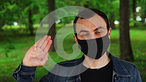 Handsome young man standing outdoors wearing medical mask and saluting by waving his hand