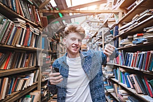 Handsome young man stands in a library with a smearphone in his hand and shows his finger down