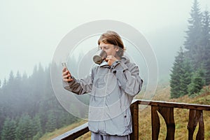 A handsome young man stands in the morning in foggy weather in the mountains on the terrace of a country house, drinking hot tea