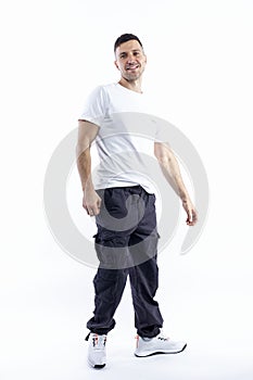 A handsome young man stands tall. A smiling brunette guy in a white T-shirt, dark trousers and sneakers. Positive and optimistic.