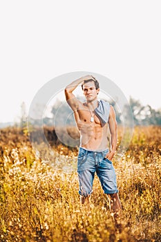 A handsome young man with a strong muscular torso in an unbuttoned shirt is standing on a meadow in nature outside city