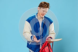 a handsome young man student with red hair stands in a fashionable jacket sadly spreading his arms holding a backpack