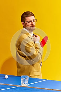 Handsome young man in stylish clothes playing tablet tennis, looking on empty whiskey glass against bright yellow