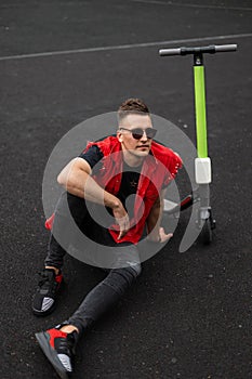 Handsome young man in sunglasses in a stylish red denim vest in black vintage jeans in sneakers sitting on the pavement