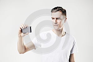 Handsome young man taking picture by mobile phone, isolated on white background, wearing white t-shirt.