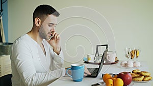 Handsome young man talking phone and using laptop computer sitting in the kitchen after breakfast in the morning