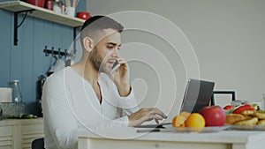 Handsome young man talking phone and using laptop computer sitting in the kitchen after breakfast in the morning