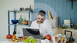 Handsome young man talking phone and using laptop computer sitting in the kitchen after breakfast in the morning