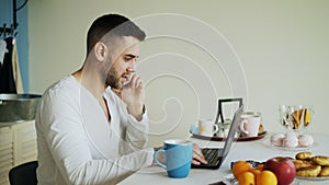 Handsome young man talking phone and using laptop computer sitting in the kitchen after breakfast in the morning