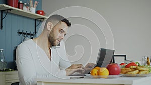 Handsome young man talking phone and using laptop computer sitting in the kitchen after breakfast in the morning
