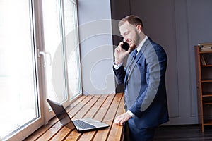 Handsome young man talking on smart phone and using laptop while working. Good business talk
