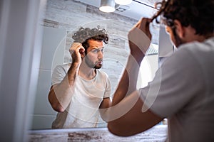 Handsome young man touching his hair with hand and grooming in bathroom at home. White metrosexual man worried for hair loss and