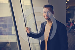 Handsome young man, transit passenger, traveler, smiling at camera, standing near panoramic windows overlooking runway in airport