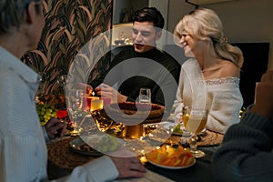 Handsome young man using match lighting candles sitting at holiday table with loving family in dark living room with