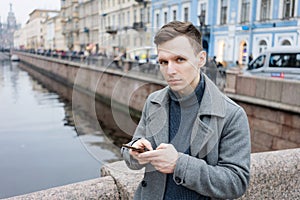 Handsome young man using mobile phone, wearing an elegant gray coat, stands