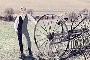 A handsome young man in a waistcoat, black trousers and a white shirt is standing next to a haymaking machine in the field. He loo