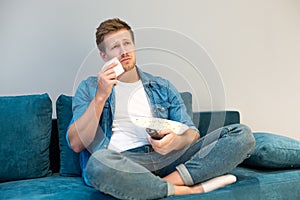 Handsome young man watching melodrama eating popcorn crying on the sofa at home