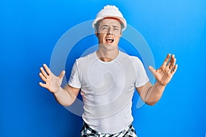 Handsome young man wearing builder uniform and hardhat crazy and mad shouting and yelling with aggressive expression and arms