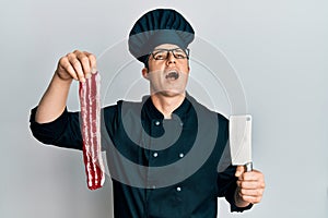 Handsome young man wearing chef uniform holding bacon and knife angry and mad screaming frustrated and furious, shouting with