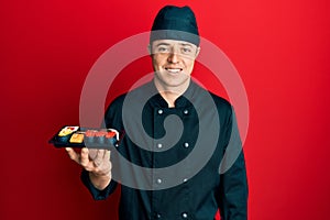 Handsome young man wearing chef uniform holding plate of sushi looking positive and happy standing and smiling with a confident