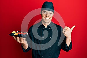 Handsome young man wearing chef uniform holding plate of sushi pointing thumb up to the side smiling happy with open mouth