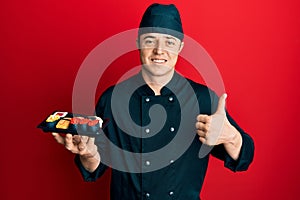 Handsome young man wearing chef uniform holding plate of sushi smiling happy and positive, thumb up doing excellent and approval